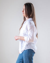 A woman with long brown hair stands in profile against a plain light background, wearing the LLEA BLUE Alix Top in White Linen and blue jeans.