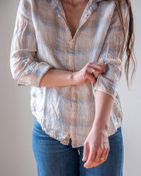 A person in blue jeans wears the LLEA BLUE Dorie Top in White, Blue, & Wheat Linen Plaid. They roll up their left sleeve, with long brown hair over one shoulder and a gold ring on their right hand. The background is plain and neutral.