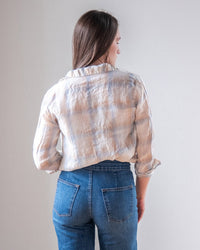 A woman with long brown hair stands facing away, wearing the LLEA BLUE Dorie Top in White, Blue, & Wheat Linen Plaid and high-waisted blue jeans against a plain light background.