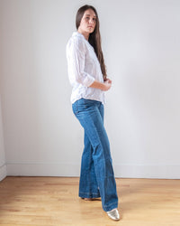 A woman with long brown hair stands indoors against a white wall, wearing the LLEA BLUE Dorie Top in White Linen, blue flared jeans, and metallic pointed-toe shoes on a light wooden floor.