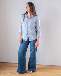 A woman stands indoors against a white wall, wearing the LLEA BLUE Dorie Top in Aqua Chambray Linen, blue wide-leg jeans, and gold shoes. She looks to her left with a slight smile.