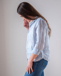 A woman with long brown hair, wearing the LLEA BLUE Dorie Top in Aqua Chambray Linen and jeans, stands in profile against a plain background, gazing down thoughtfully with her hand near her face.