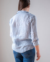 A person with long brown hair, facing away from the camera against a plain background, wears blue jeans and an LLEA BLUE Dorie Top in Aqua Chambray Linen.