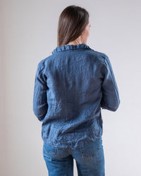 A person with long brown hair, wearing LLEA BLUE's Dorie Top in Moonlit Ocean Linen and jeans, stands facing away from the camera against a plain light background.