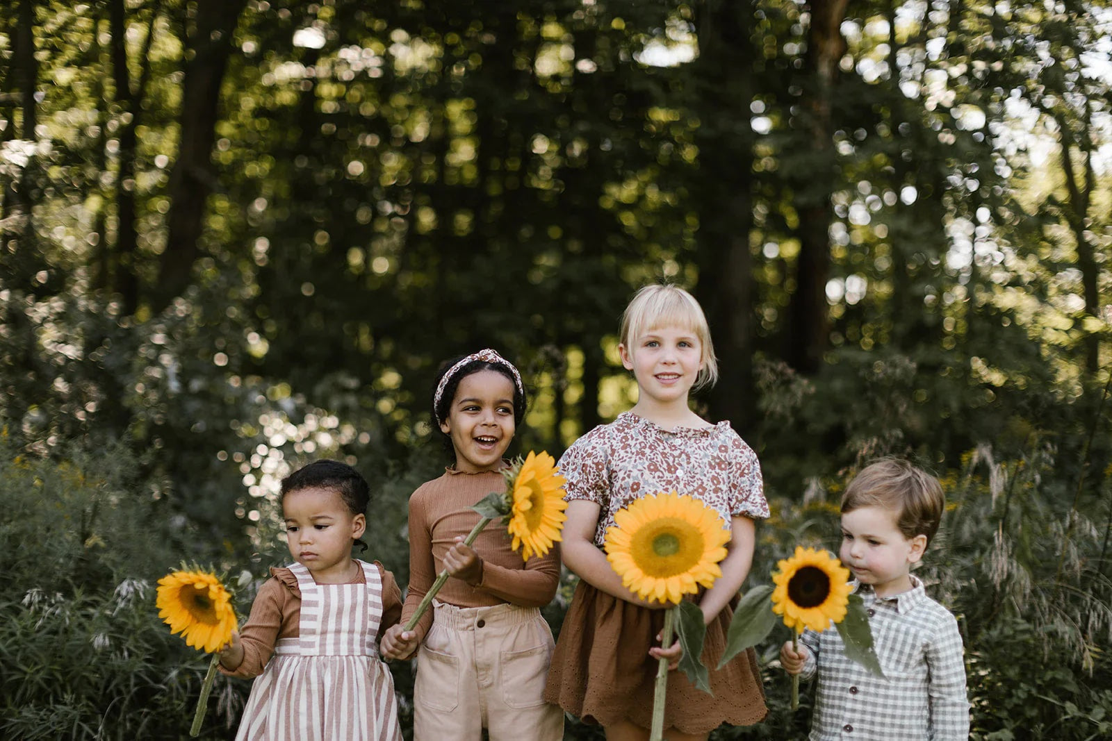 Four children standing in a forest, each holding a sunflower.
