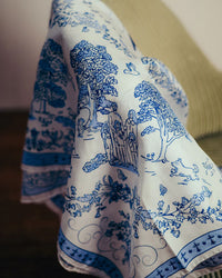 A Maison Lecomte Flament Toile de Jouy 100% Silk scarf in navy, featuring line drawings of trees, people, a dog, and florals, is draped over a green-striped chair, showcasing refined Italian artistry.