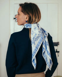 A woman with light brown hair, in a navy sweater and beige pants, stands with her back to the camera. She wears Maison Lecomte Flament’s Toile de Jouy 100% Silk in Navy scarf around her neck and faces a white door.