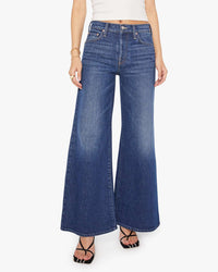 A woman, visible only from the waist down, wears the Mother Lil Tomcat Roller Skimp in Perfectly Imperfect—high-waisted wide-leg jeans made from premium denim—paired with a white sleeveless top and black strappy sandals.