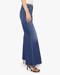 A person in a white top, Mother Lil Tomcat Roller Skimp in Perfectly Imperfect jeans, black heeled sandals, and gold bracelets stands against a plain white background.