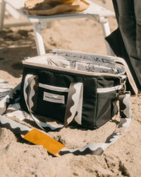 The Premium Cooler Bag - Rivie Black by Business & Pleasure Co, featuring patterned straps and a vintage look, sits open on sandy beach near a white chair.