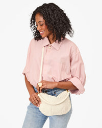 A woman in a loose pink button-up shirt and light blue jeans holds the Clare V. Petit Moyen Messenger - Woven Checker in Cream crossbody bag, standing against a plain white background.