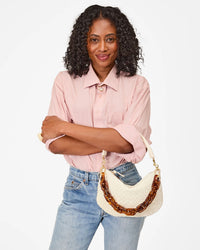 A woman with curly hair wears a light pink shirt and blue jeans, holding the Clare V. Petit Moyen Messenger - Woven Checker in Cream, featuring a brown woven leather chain strap.