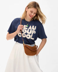 A smiling blonde woman wears a navy BEAUCOUP COOL shirt, white skirt, and holds the Clare V. Petit Moyen Messenger in Smooth Nubuck Hazelnut against a plain white background.