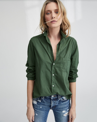 A woman with wavy blonde hair wears the Frank & Eileen Eileen Relaxed Button Up Shirt in army green, styled with blue distressed jeans, standing against a light, neutral background.