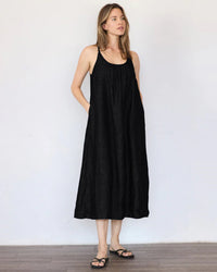 A woman stands indoors against a plain wall, wearing the It is well LA Linen Tank Dress in Black—a sleeveless, loose-fitting A-line dress with pockets—paired with black sandals.