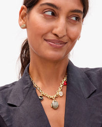 A woman in a dark blazer smiles slightly, wearing a gold NON earring and a chunky vintage gold-plated brass chain with several charms, including the Clare V. Stripe Locket in Vintage Gold/Black and a red accent.