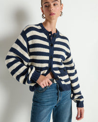 Against a white wall, a person with short hair wears Nation LTD’s Tessa Cardigan in Marine Stripe—an oversized navy and white striped look—paired with blue jeans and silver hoop earrings.