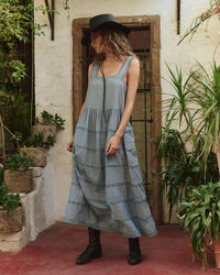 A person in the Greats Reverie Dress in Pale Blue, featuring a loose fit and tiered skirt, pairs it with a black hat and boots while standing on a red floor near green plants and a wooden door.