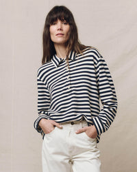 A person wearing the Rugby Sweatshirt in Navy Sailor Stripe by the Great, along with white pants, stands confidently against a plain backdrop.