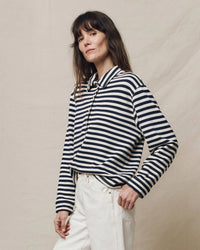 A person wearing the Greats Navy Sailor Stripe Rugby Sweatshirt and white pants stands in front of a neutral backdrop.