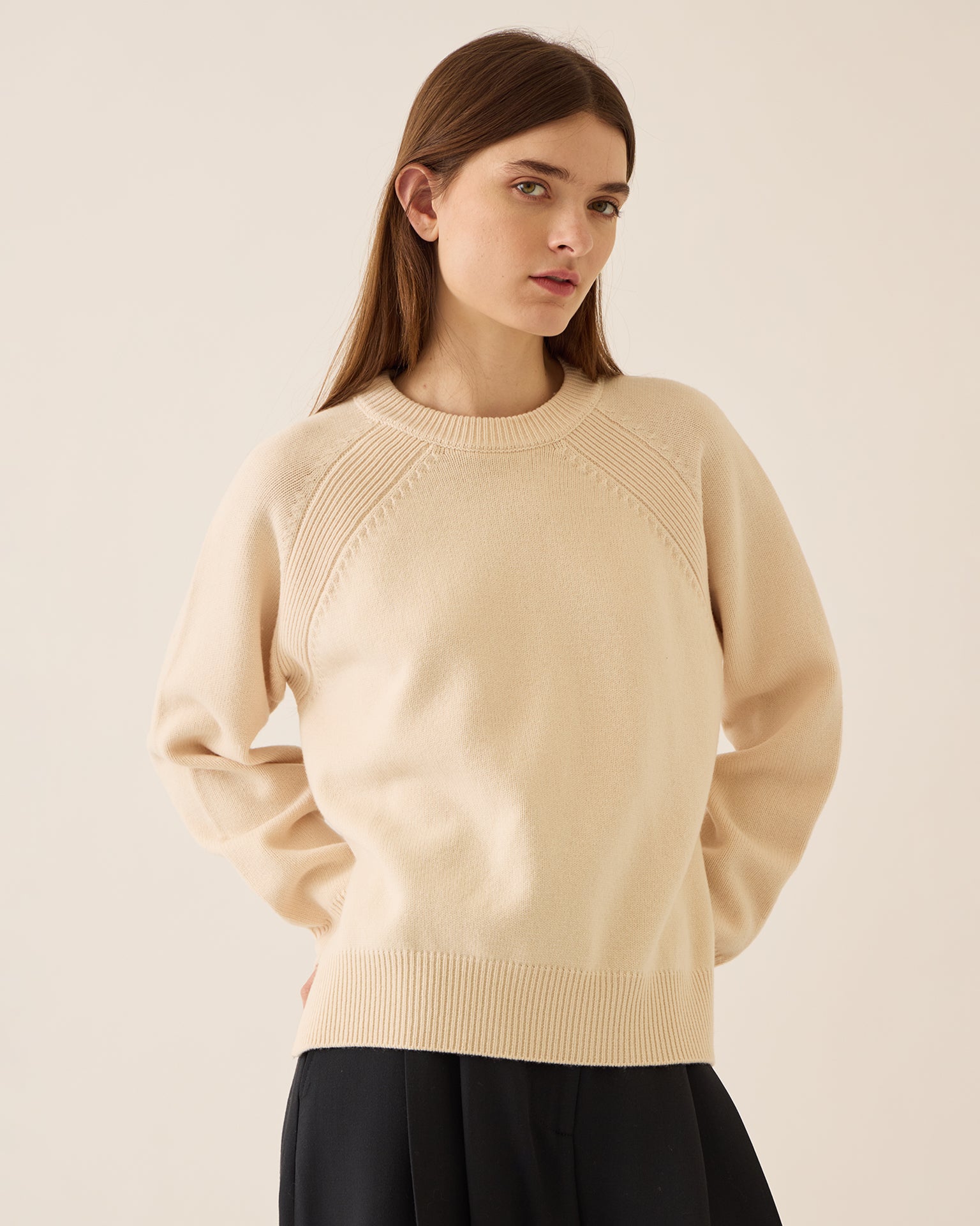 A woman with long brown hair wears the Demylee Briella Merino Wool Sweater in Off White and a black skirt, standing against a plain light background and gazing slightly to the side with a neutral expression.