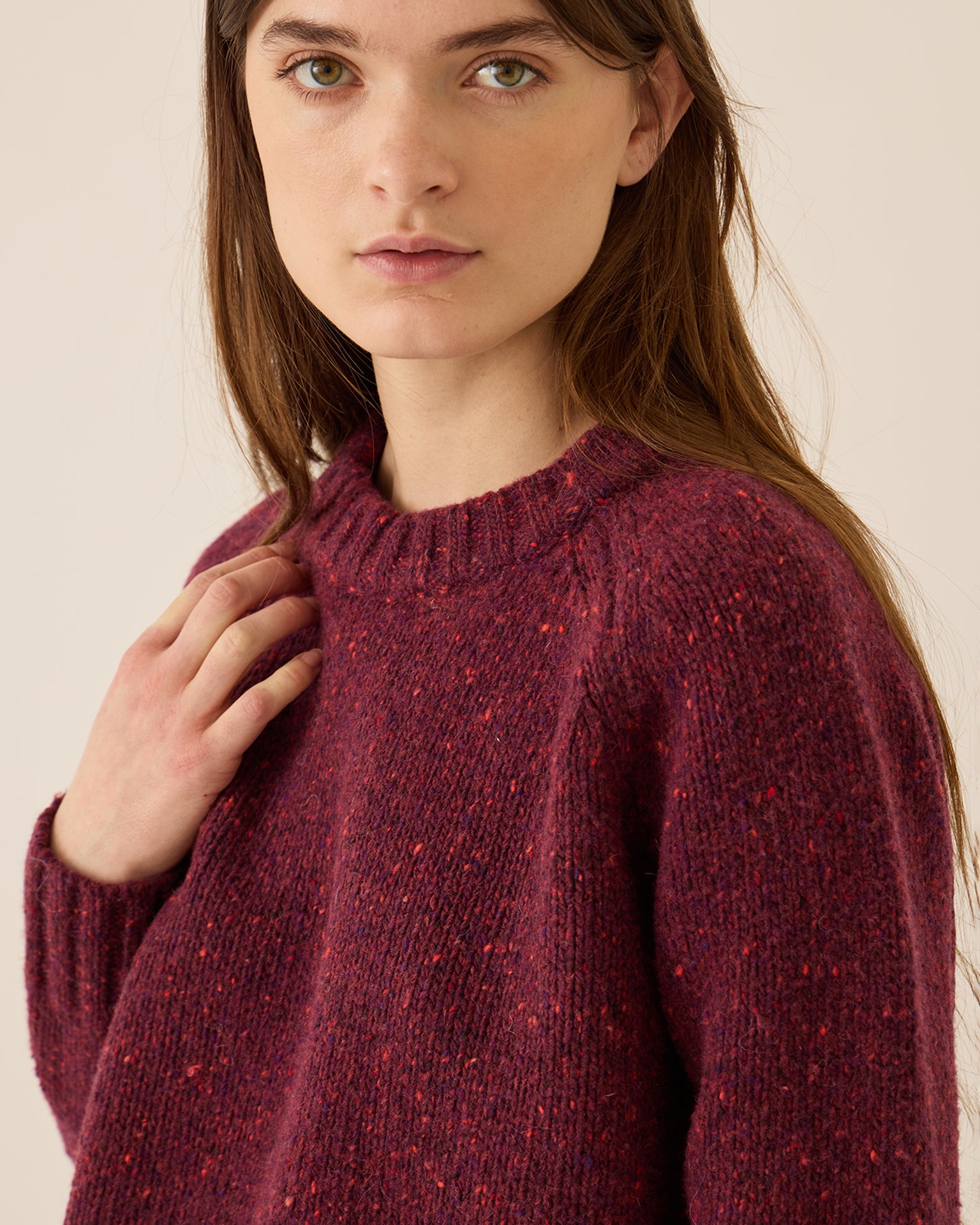 A young woman with long brown hair models the Demylee Emberly Speckled Wool Sweater in Aubergine, featuring a relaxed fit. She gazes neutrally at the camera, her hand near her collarbone, set against a plain light background.