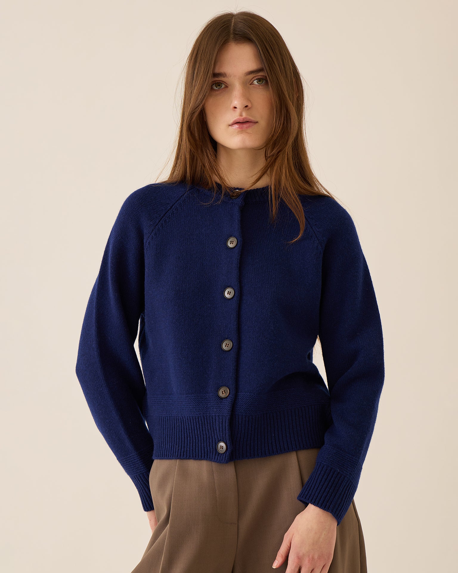 A woman with long brown hair is wearing the Demylee Frances Merino Wool Cardigan in Sapphire and light brown trousers, standing against a plain beige background.