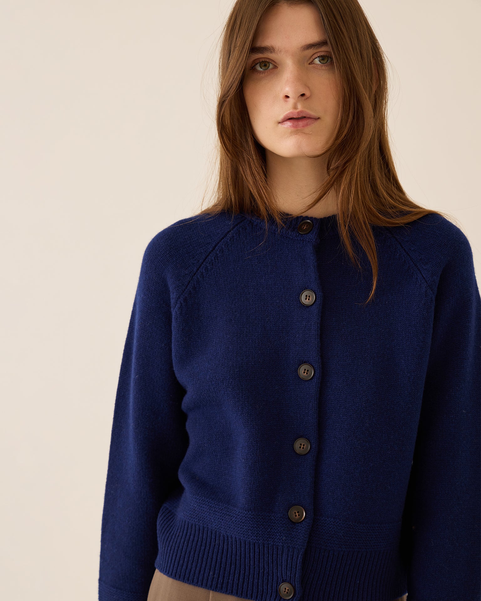 A woman with long brown hair wears the Demylee Frances Merino Wool Cardigan in Sapphire, featuring a crewneck and long sleeves, standing against a plain light beige background and looking directly at the camera.