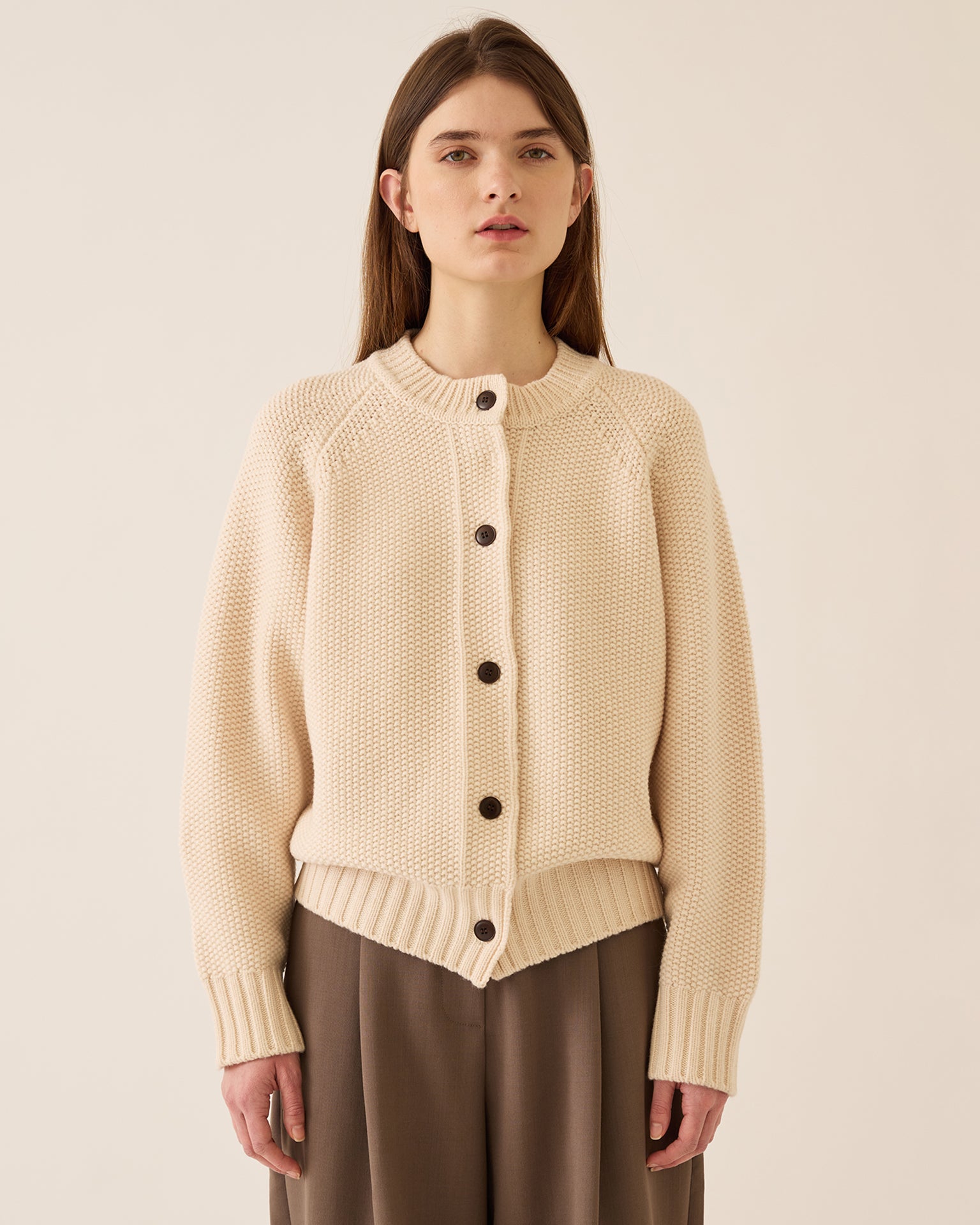 A woman with straight brown hair wears the Demylee Davi Merino Wool Cardigan in Off White, featuring dark buttons and wide ribbed cuffs, paired with loose brown pants, standing against a plain light background.