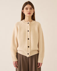 A woman with straight brown hair wears the Demylee Davi Merino Wool Cardigan in Off White, featuring dark buttons and wide ribbed cuffs, paired with loose brown pants, standing against a plain light background.