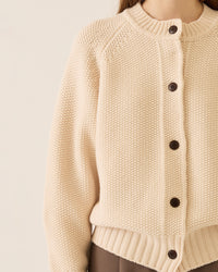 A person wears the Demylee Davi Merino Wool Cardigan in Off White with dark buttons, paired with brown pants. The cropped image shows only their lower face and torso, from shoulders to mid-thigh.