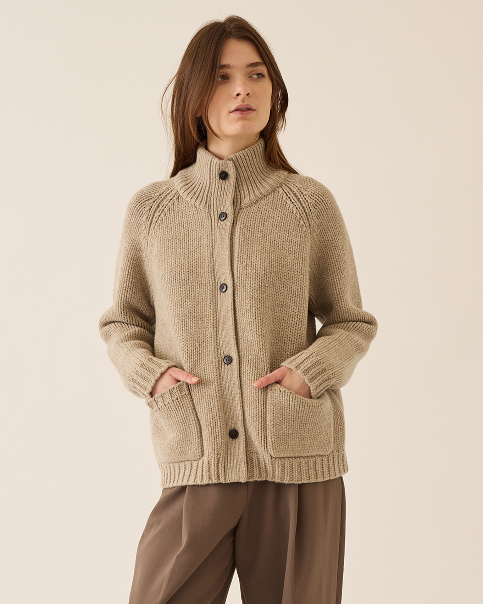 A woman stands against a plain light background wearing Demylee's Kamila Soft Chunky Wool Cardigan in Oatmeal, featuring a high collar and front pockets, paired with brown pants.