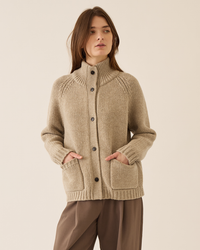 A woman stands against a plain light background wearing Demylee's Kamila Soft Chunky Wool Cardigan in Oatmeal, featuring a high collar and front pockets, paired with brown pants.