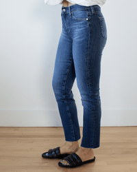 A person wearing AG Jeans Mari Crop in 14Ys Collector, a white long-sleeve shirt, and black slide sandals stands indoors on a wooden floor against a plain white wall.