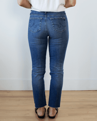 A person stands indoors facing away, wearing AG Jeans Mari Crop in 14Ys Collector denim with a white top and black sandals on a wooden floor against a white wall.