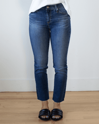 A person stands on a wooden floor against a plain light-colored wall, wearing AG Jeans' Mari Crop in 14Ys Collector denim, paired with a white top and black slide sandals.