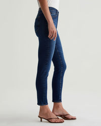 A person wearing AG Jeans' Prima Ankle in Catalogue—dark blue, mid-rise cigarette leg—paired with a white top and brown heeled sandals, stands against a plain light background, shown from the shoulders down in side view.