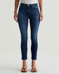 A person wearing a white top, brown sandals, and AG Jeans' Prima Ankle in Catalogue—a dark blue, cigarette-leg, mid-rise jean with classic five-pocket styling—stands against a plain light background. The person’s face is not shown.