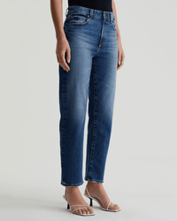 A person wears AG Jeans' Rian High-Rise Straight in Lake Como, featuring a vintage-inspired faded wash, paired with a black top and white open-toe heeled sandals, standing against a plain light background.