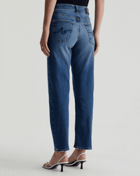A person wearing AG Jeans' Rian High-Rise Straight in Lake Como and white heeled sandals, shown from the back against a plain light background, captures a vintage-inspired fit with effortless style.