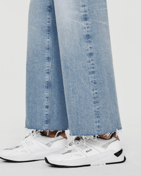 A person wears AG Jeans’ Saige Wide Leg Crop in Chateau—light blue, high-rise, cropped wide-leg jeans with frayed hems—and white BOSS athletic sneakers, standing against a plain background.