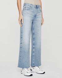A person wears the Saige Wide Leg Crop in Chateau by AG Jeans, featuring light blue, high-rise, cropped wide-leg jeans, paired with white athletic sneakers against a plain light background.