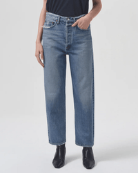 A person wearing AGOLDE 90s Crop Mid Rise Loose Straight in Hooked jeans and black ankle boots stands with one hand in their pocket against a plain light background.