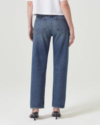 A person in a white shirt, 90s Jean in Imagine by AGOLDE (high-waisted blue jeans), and black heeled sandals stands facing away, showing the back view of the jeans against a plain light background.