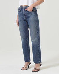A person wearing AGOLDE’s 90s Jean in Imagine—high-waisted, straight-leg blue jeans—paired with a white cropped top and black heeled sandals stands against a plain white background. Their head is not shown.