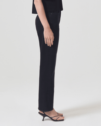 A person stands in profile against a white background, wearing AGOLDE 90s Pinch Waist in Crushed jeans, a black top, and black open-toe heeled sandals.