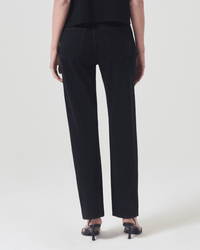A person stands facing away, wearing AGOLDE 90s Pinch Waist jeans in Crushed, a black top, and black heeled sandals against a plain light background.