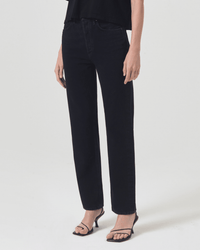 Against a plain white background, only the lower half of a person is visible. They wear AGOLDE 90s Pinch Waist jeans in Crushed, paired with a black top and black strappy heeled sandals.