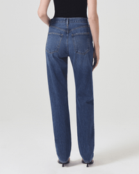 A person wearing a black sleeveless top and AGOLDE 90s Pinch Waist Long in Control jeans stands facing away, showcasing the back pockets and seam details against a plain, light-colored background.