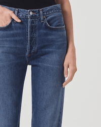 A person wears AGOLDE 90s Pinch Waist Long in Control jeans with a black top, one hand in their pocket and the other at their side, shown from waist to mid-thigh against a plain background.